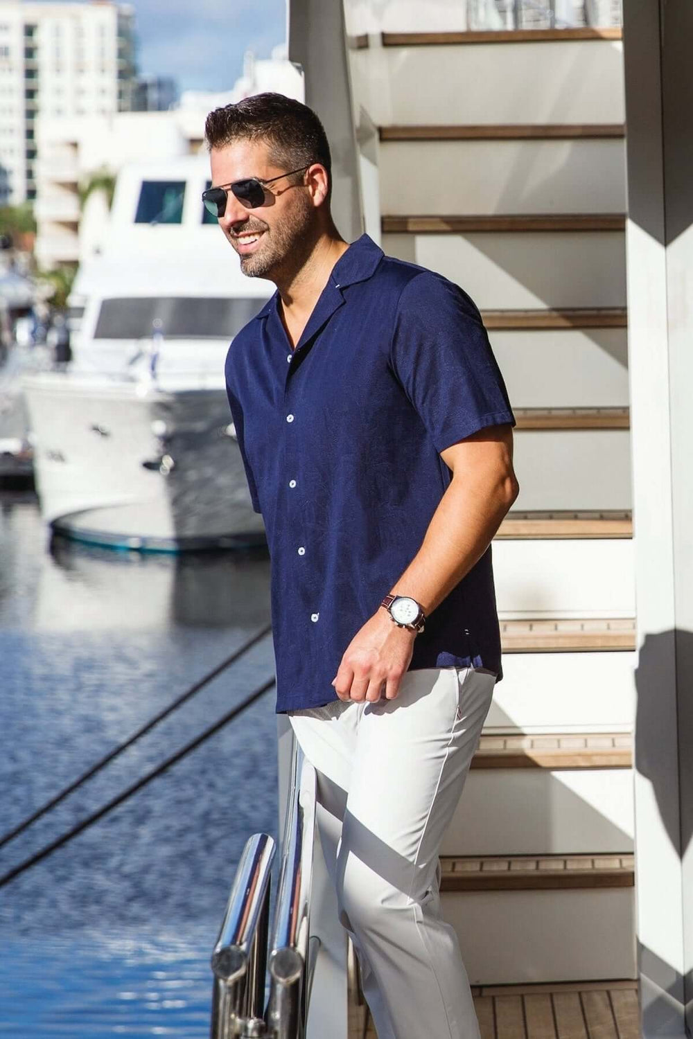 Homme portant une chemise à manches courtes bleu marine, lunettes de soleil, marchant au bord d'un quai.