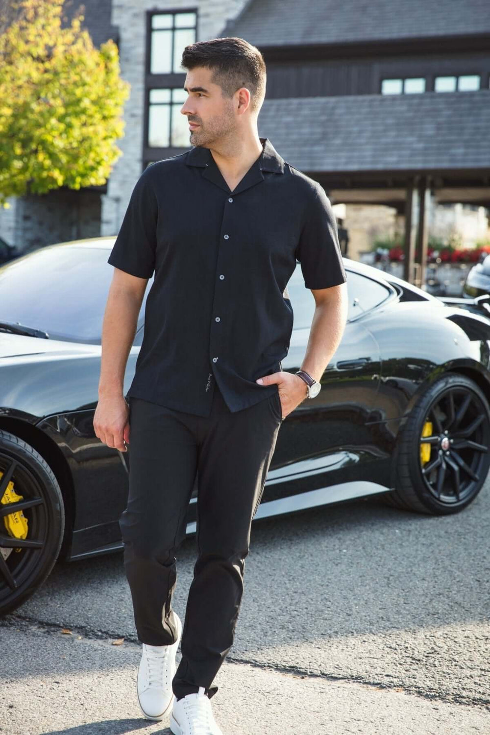 Homme portant une chemise noire à col cubain et un pantalon noir, posé près d'une voiture de sport élégante.