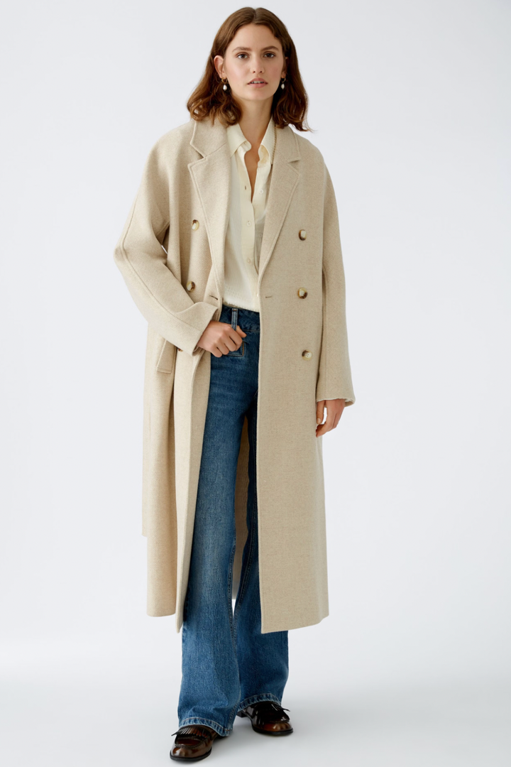 Femme en manteau beige sur chemise blanche et jeans évasés, alliant confort et élégance dans une pose moderne.