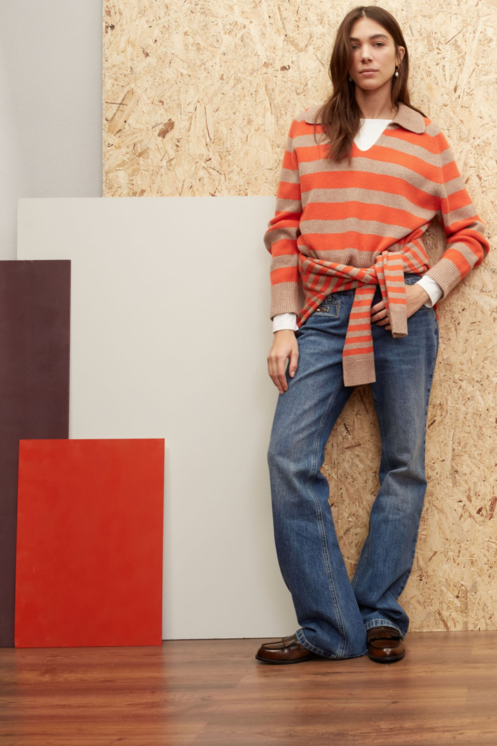 Femme portant un pull rayé orange et un jean évasé dans un décor moderne avec des fonds colorés.