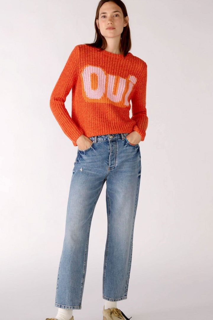 Femme portant un pull orange avec 'OUi' en lettres blanches et un jean droit bleu. Style décontracté.