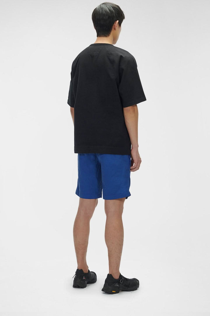 Homme de dos portant un t-shirt noir et un short bleu, avec des chaussures noires, sur fond blanc.