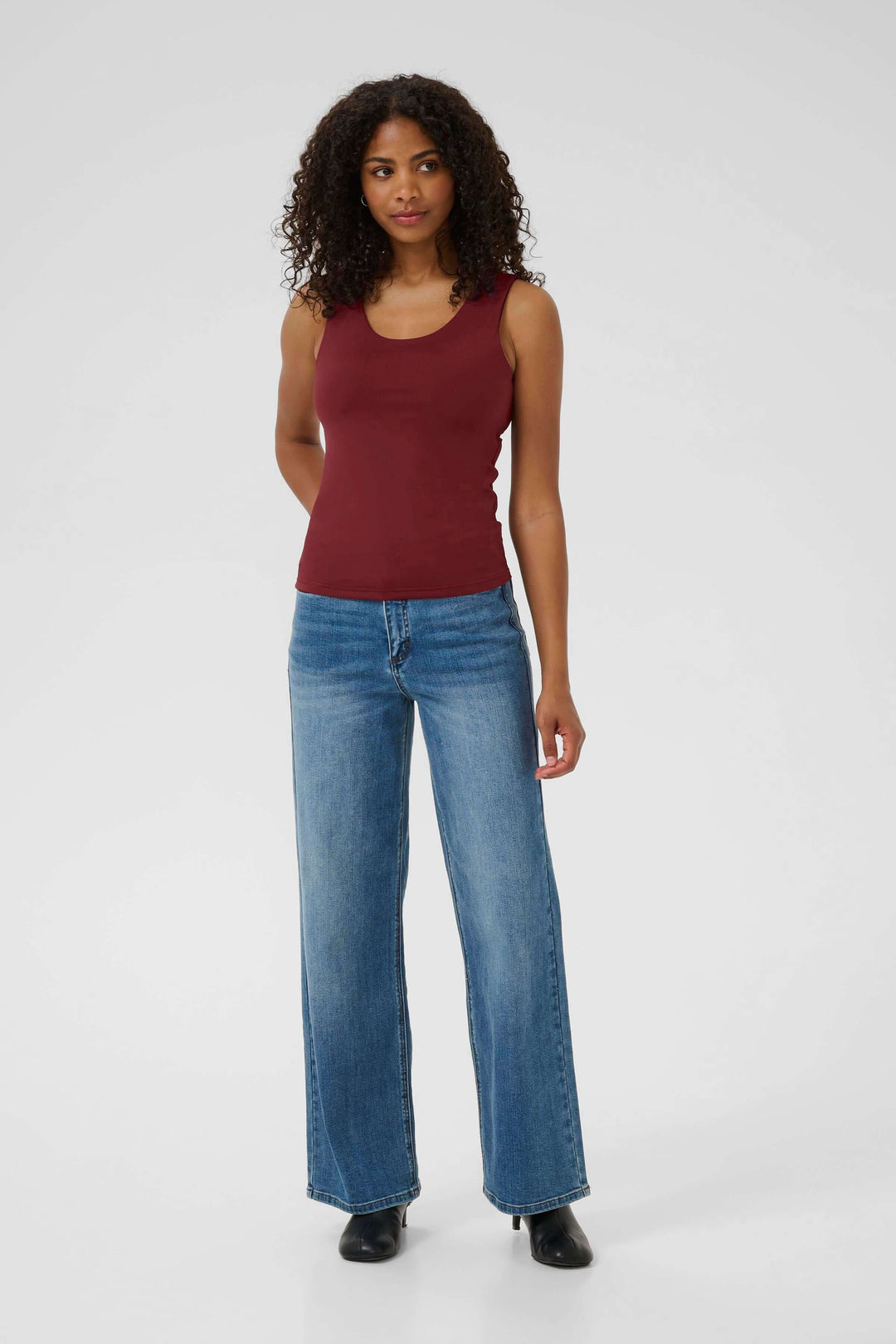 Woman wearing a burgundy tank top with blue wide-leg jeans and black shoes, standing in a neutral pose against a gray background.