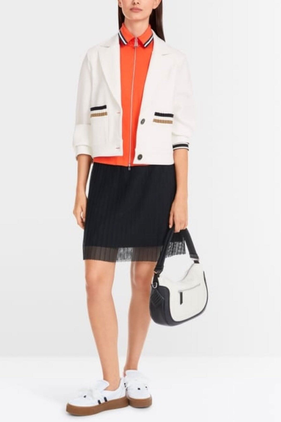 Chic woman in a white jersey blazer, orange top, and pleated black skirt, carrying a stylish handbag.