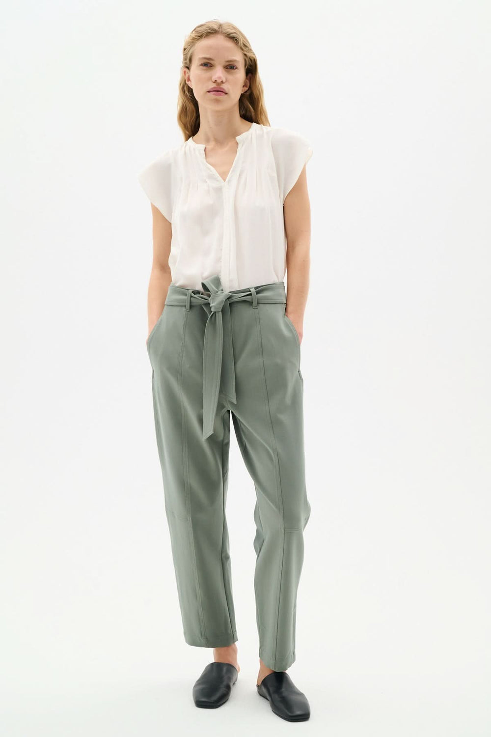 Model wearing a light blouse with pleated neckline and green high-waisted trousers, styled elegantly for a chic look.