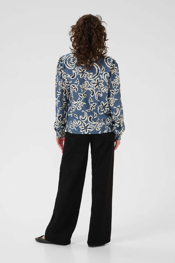 Femme de dos portant une blouse Dahlia avec manches longues et tissu ajouré, associée à un pantalon noir pour un look élégant.