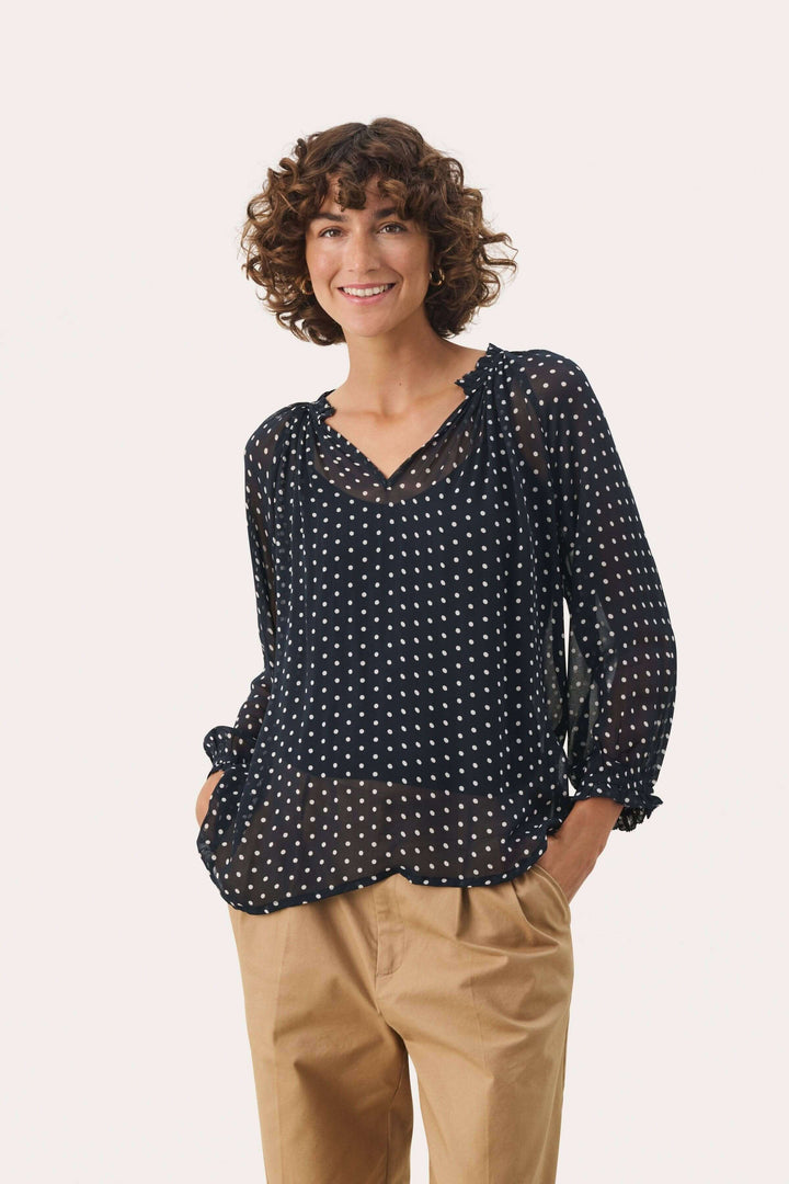 Femme portant une blouse Elsia en viscose noire à pois, avec manches 3/4 et coupe décontractée, associée à un pantalon beige.