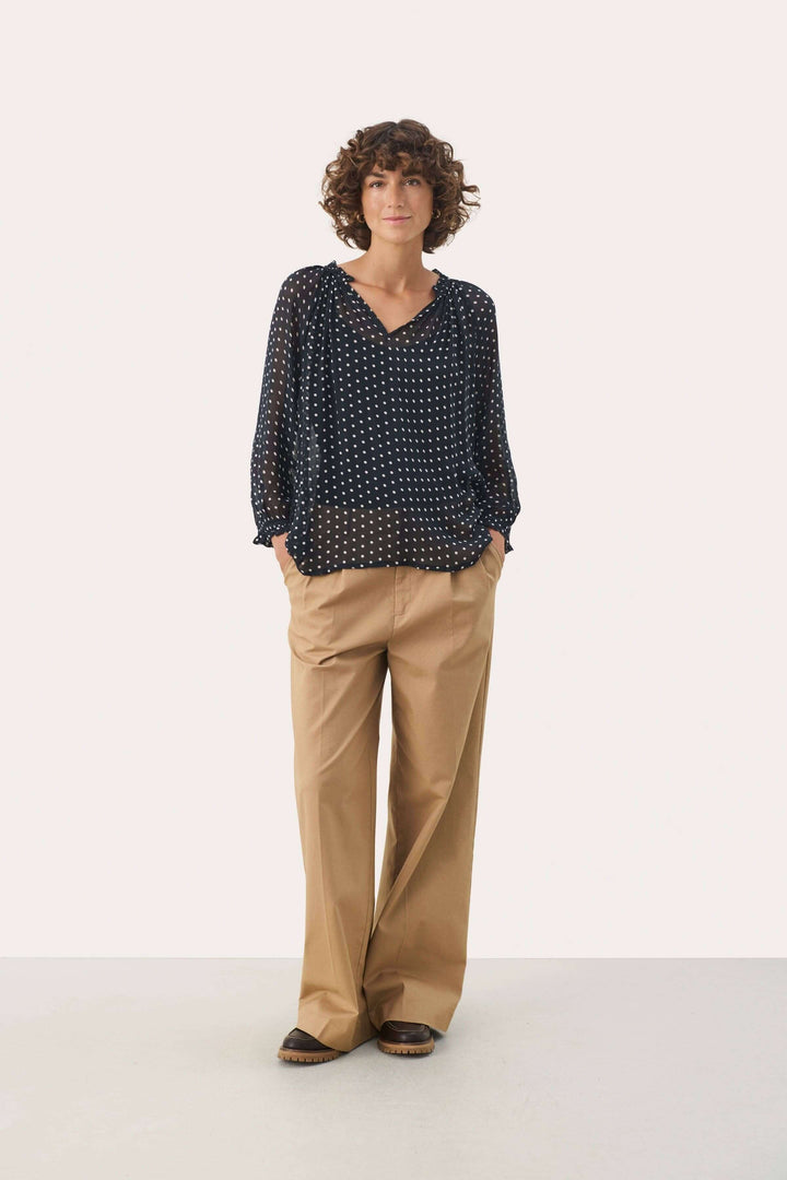 Femme portant une blouse Elsia à imprimé noir et blanc avec des manches 3/4 et un pantalon beige décontracté.