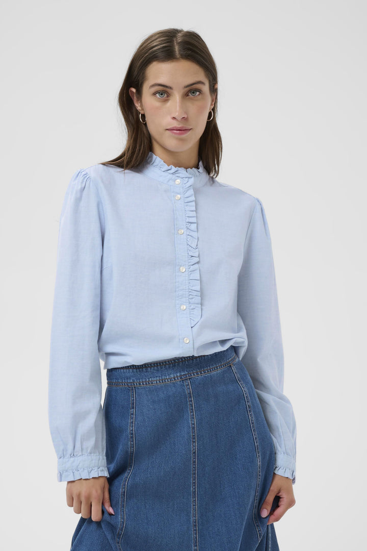 Blouse Glessia romantique avec volants délicats, portée sur une jupe denim. Coupe fluide et féminine, col volanté, boutons devant.