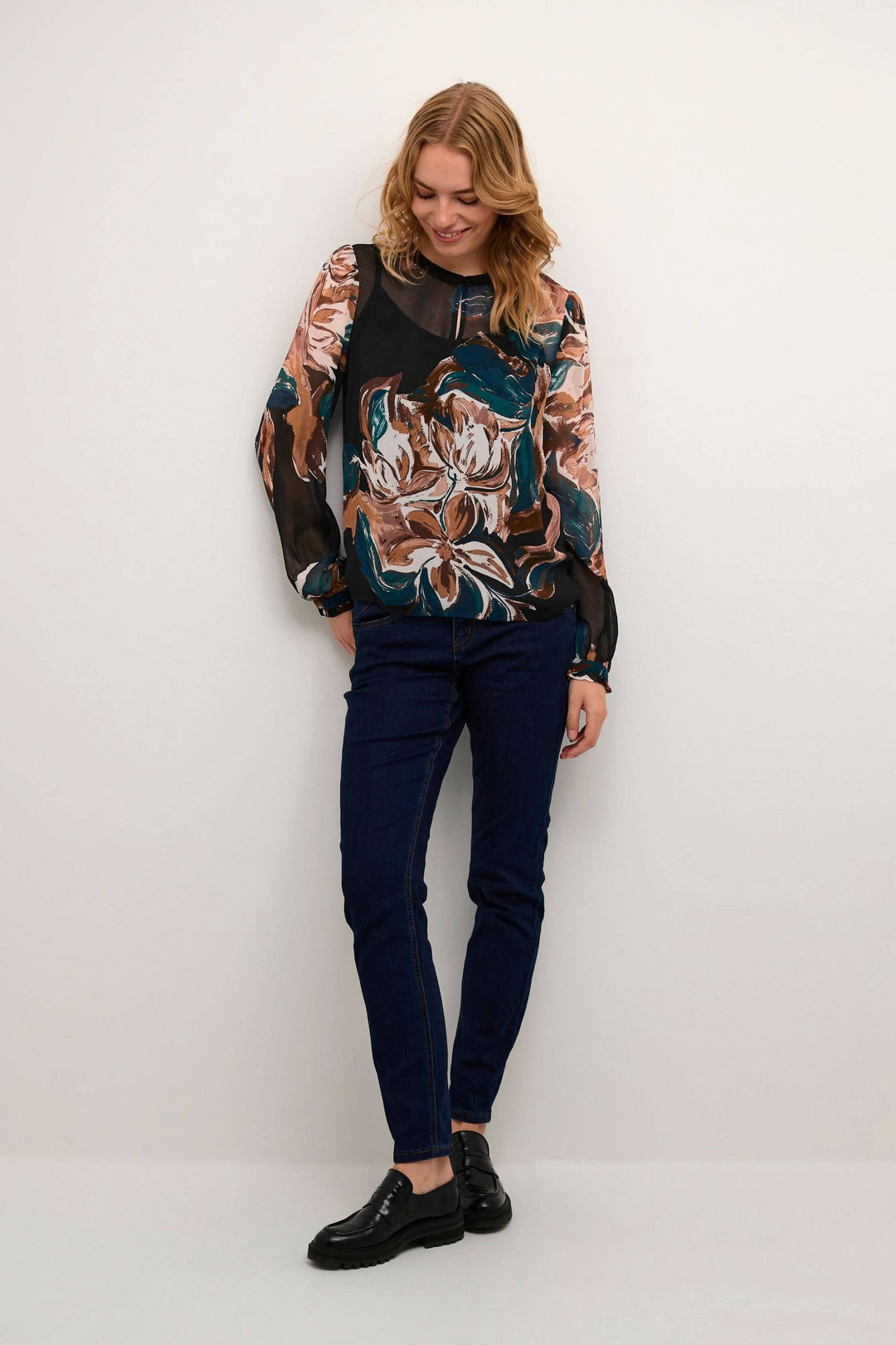 Blouse Jasmina à imprimé floral, avec encolure en V et manches longues, portée avec un pantalon bleu.