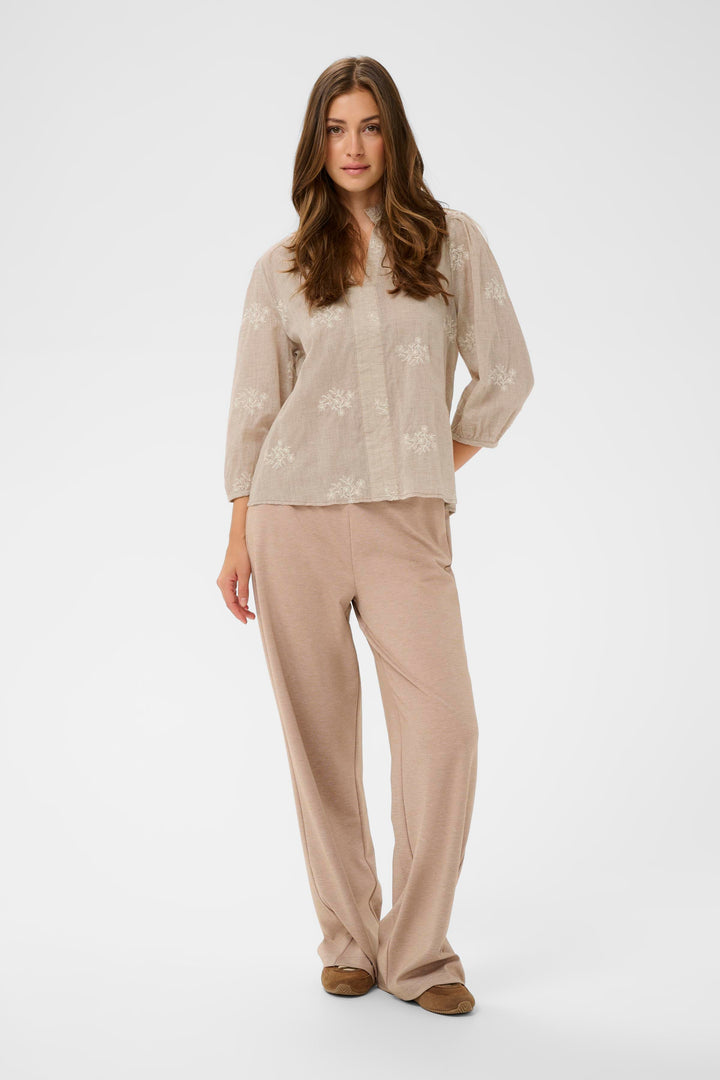 Femme portant une Blouse Korey avec encolure en V ouverte et motif discret, combinée avec un pantalon beige, mise en avant du style raffiné.