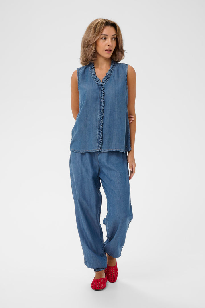 Femme portant la Blouse Molly sans manches en denim avec encolure en V et détail volanté, combinée à un pantalon assorti.