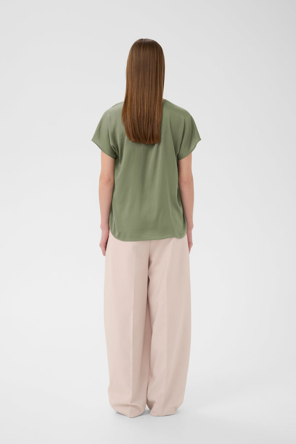 Dame portant la Blouse Rinda fluide en vert avec un pantalon beige, vue de dos, encolure en V et coupe droite élégante.