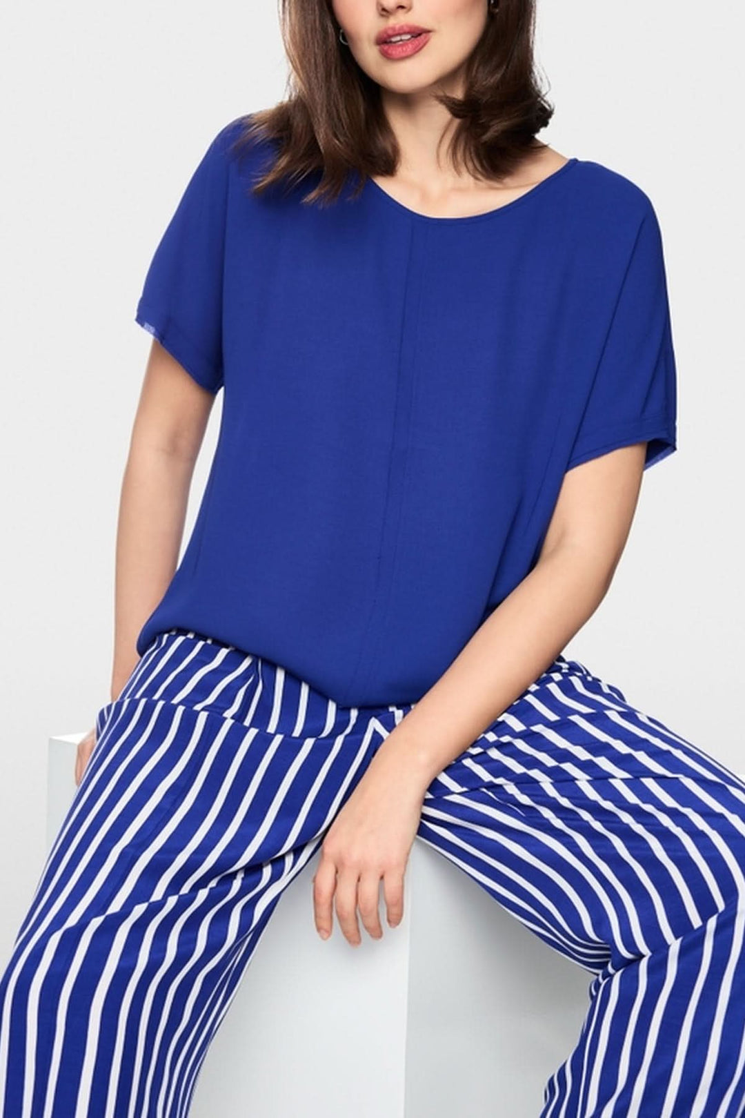 Femme portant une blouse ample en tissu fluide bleu avec un décolleté rond et manches courtes, associée à un pantalon rayé bleu et blanc.