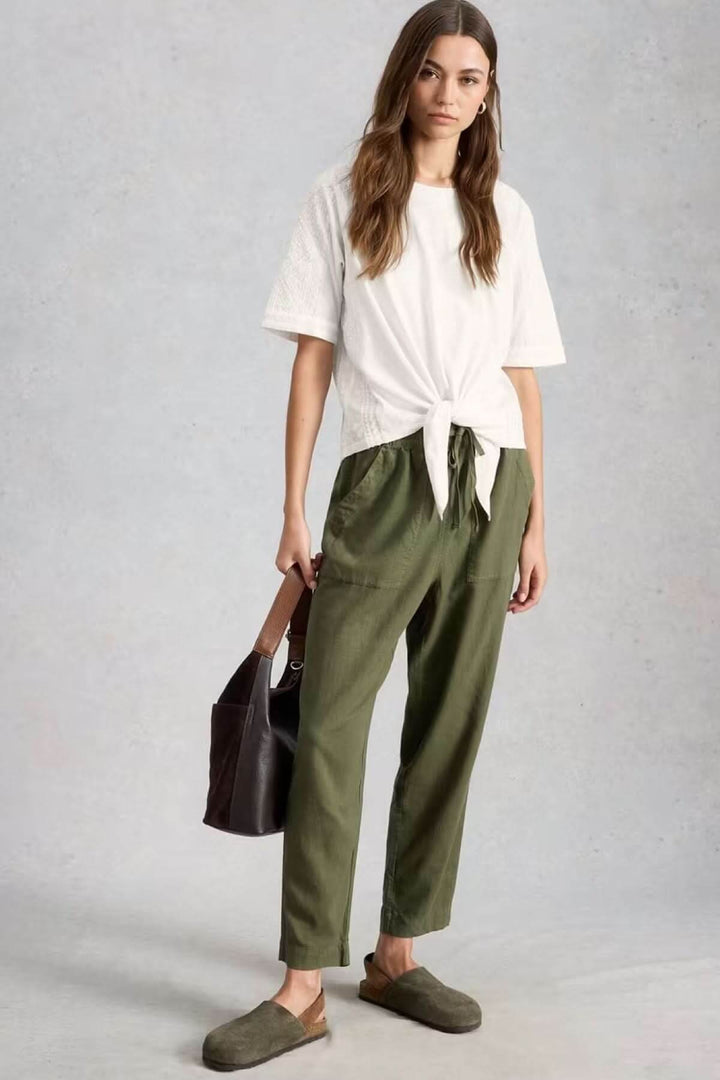 Femme portant une blouse à col rond brodé avec ourlet à nœud, tenue décontractée avec pantalon vert et mule en daim.