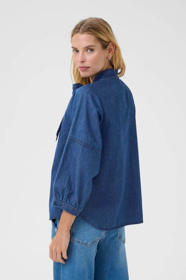 Femme portant une blouse en denim bleu, col noué, manches bouffantes, vue de dos.