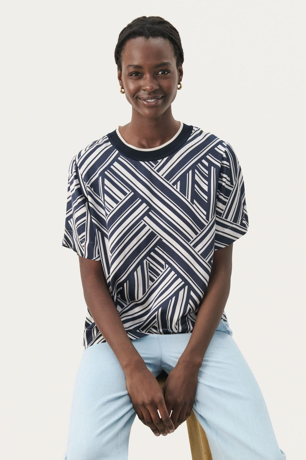 Model wearing a casual navy and white striped blouse, sitting on a stool, smiling.