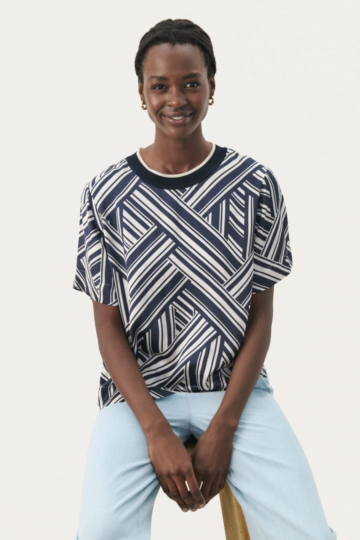 Model wearing a casual navy and white striped blouse, sitting on a stool, smiling.