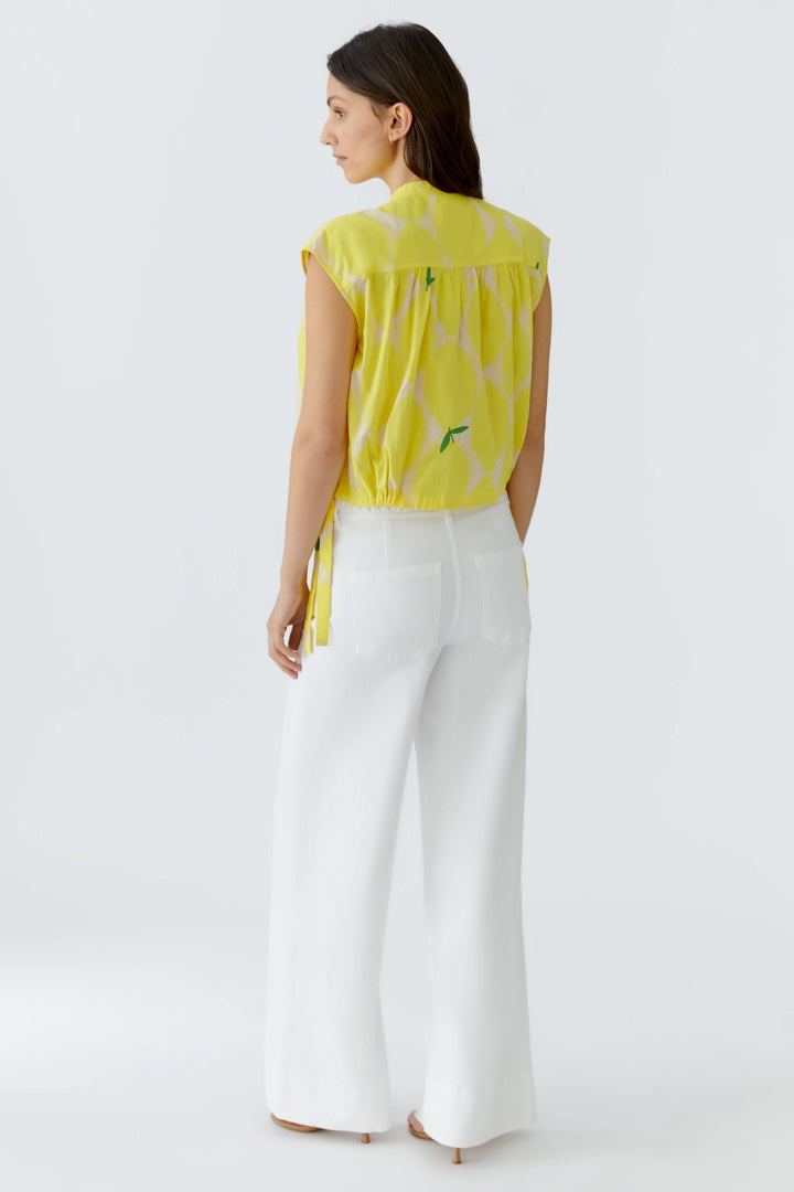 Vue arrière de la blouse fluide à col montant jaune citron, sans manches, portée avec un pantalon blanc pour un look estival chic.