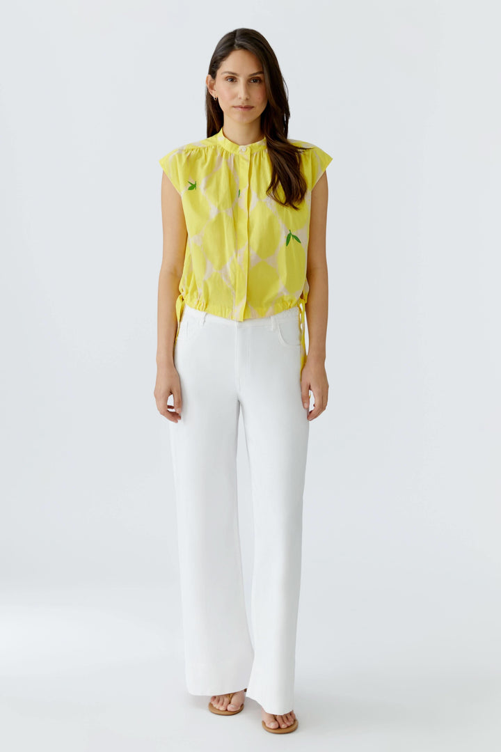 Femme portant une blouse fluide à col montant jaune avec motif, sans manches, associée à un pantalon blanc élégant, vue de face.
