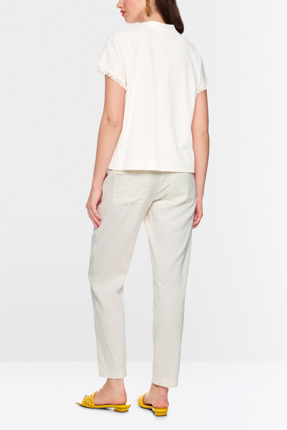 Blouse fluide à finitions effilochées avec manches courtes, vue arrière, portée avec un pantalon blanc et des sandales jaunes.