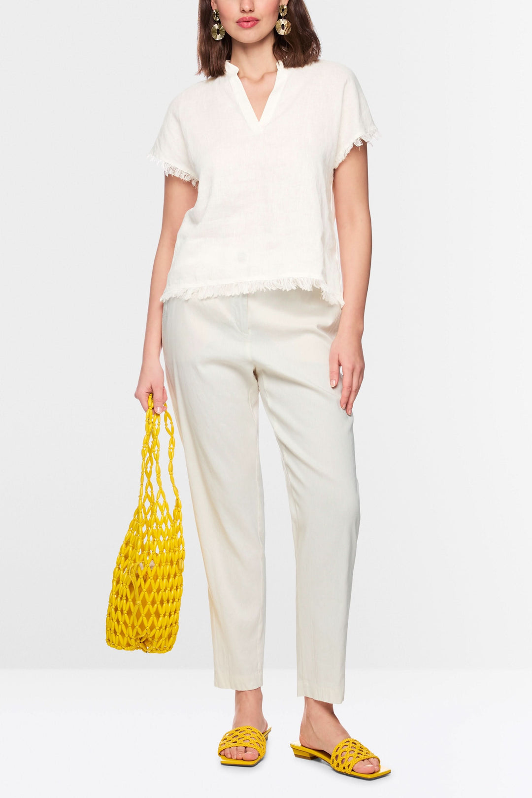 Femme portant une blouse fluide à finitions effilochées, encolure en V, manches courtes, avec un pantalon blanc et accessoires jaunes.