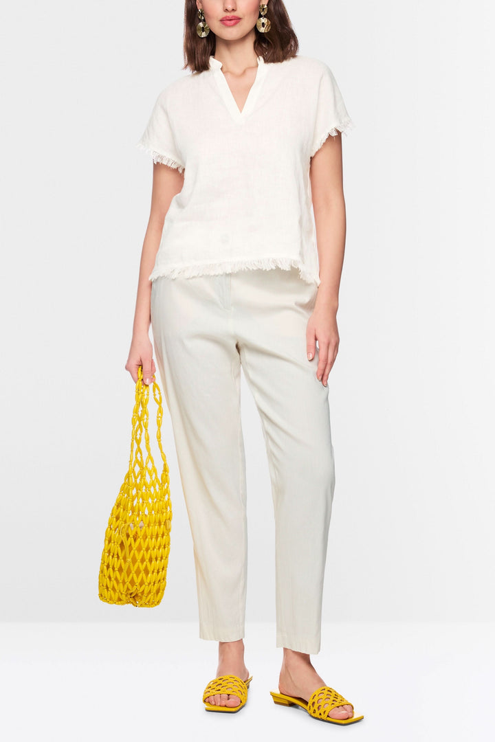 Femme portant une blouse fluide à finitions effilochées, encolure en V, manches courtes, avec un pantalon blanc et accessoires jaunes.
