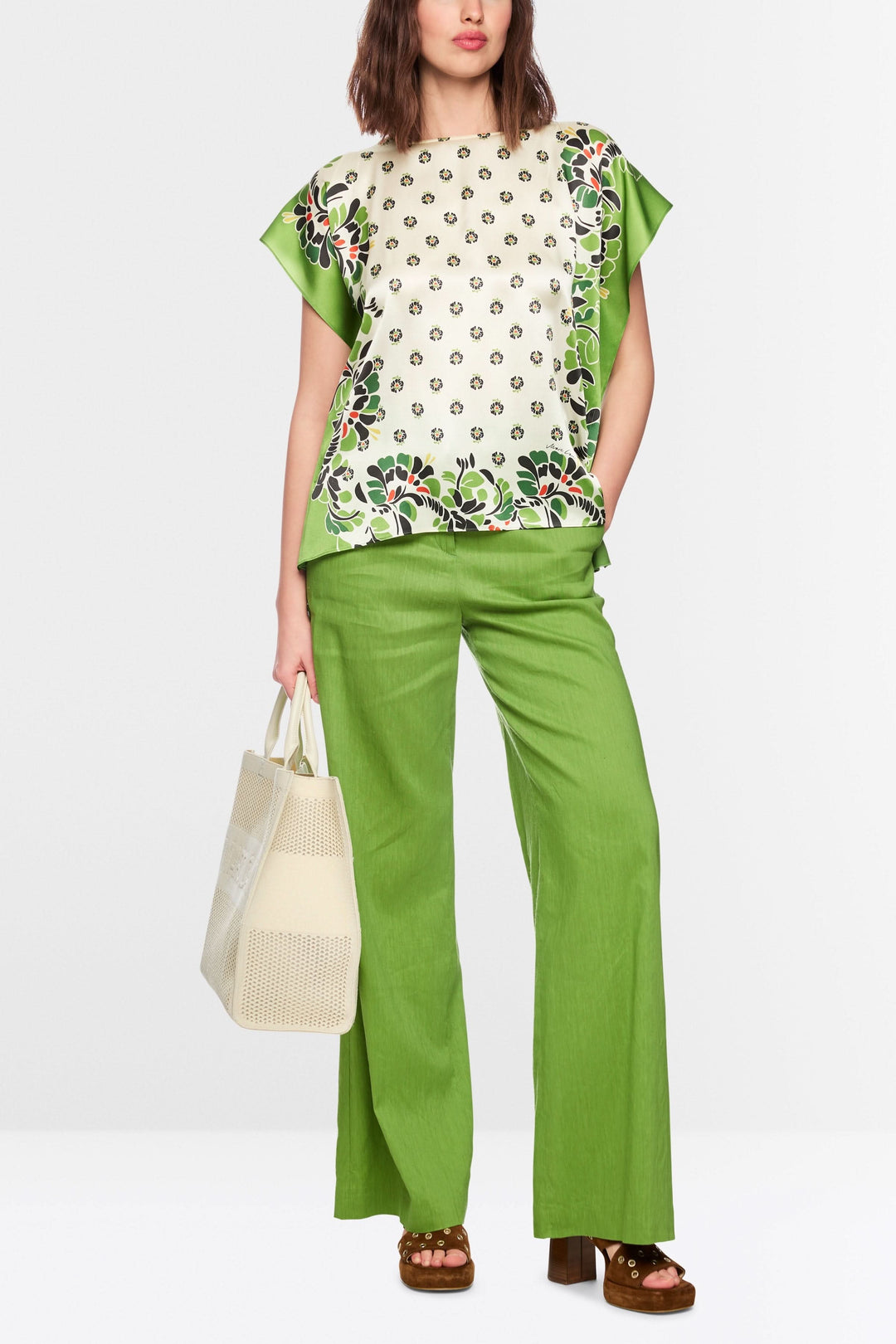 Blouse fluide imprimée à manches courtes amples, portée avec un pantalon vert, et un sac en osier, affichant un style polyvalent.