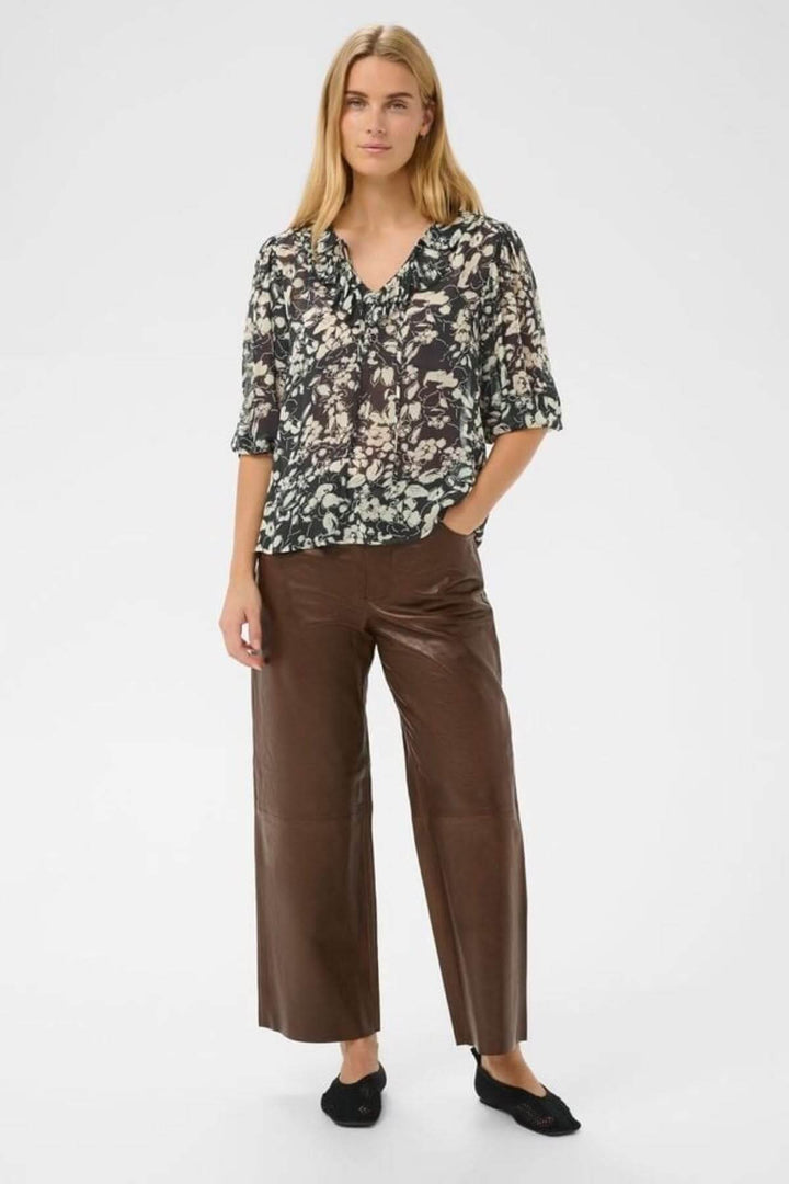 Femme portant la blouse fluide à volants Alvia avec pantalon brun, style décontracté et élégant.