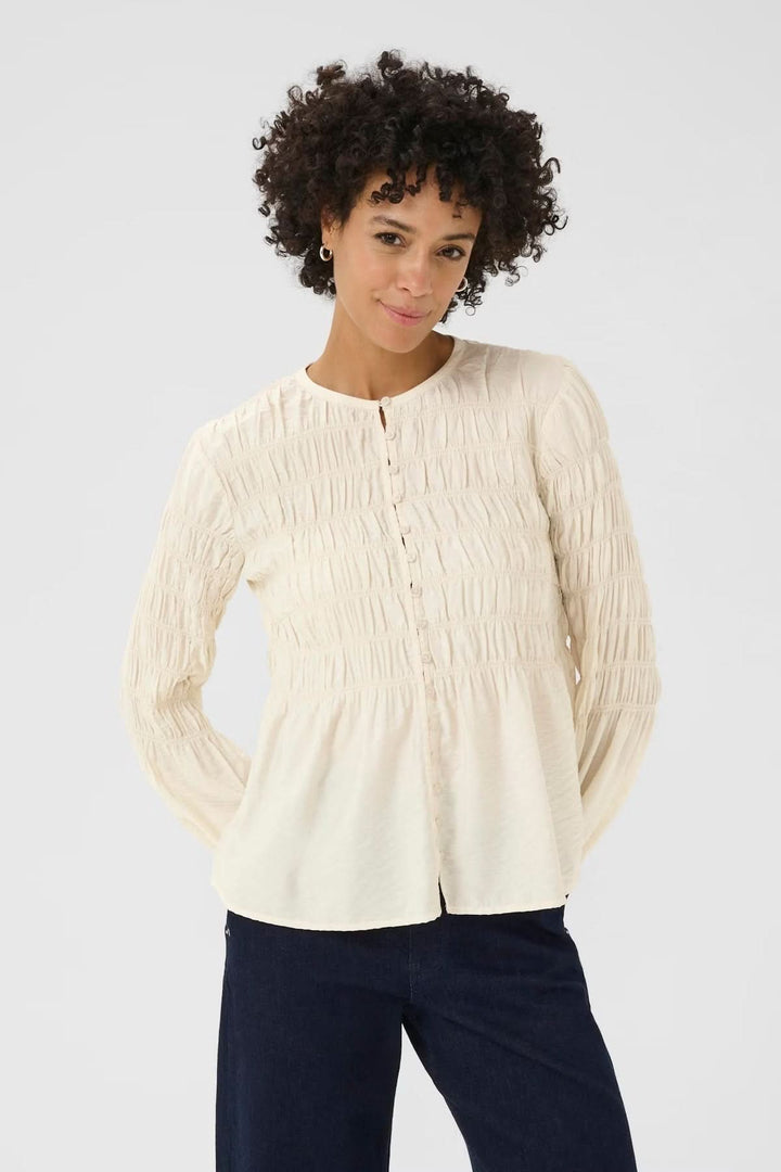 Femme portant une blouse à fronces beige avec encolure ronde et patte boutonnée, tenue casual chic, arrière-plan gris clair.