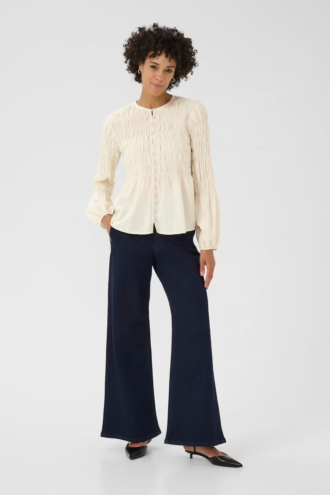 Blouse à fronces avec coupe fluide et détails texturés, encolure ronde et patte boutonnée, portée avec un pantalon ample bleu.
