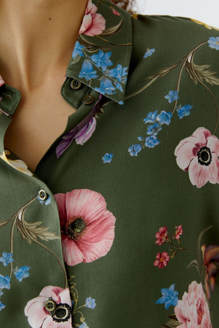 Blouse à imprimé floral fluide avec motifs de fleurs roses et bleues sur fond vert, détails du col chemise classique.