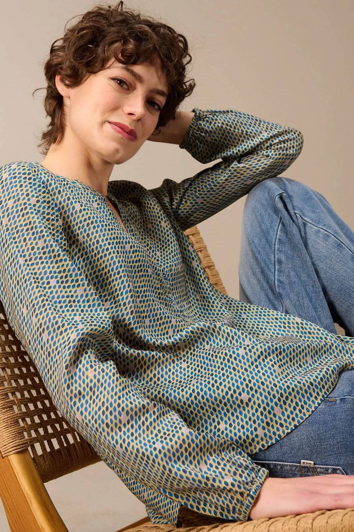 Blouse imprimée à manches longues, femme assise sur une chaise en rotin portant un jean, mettant en valeur l'imprimé graphique subtil.