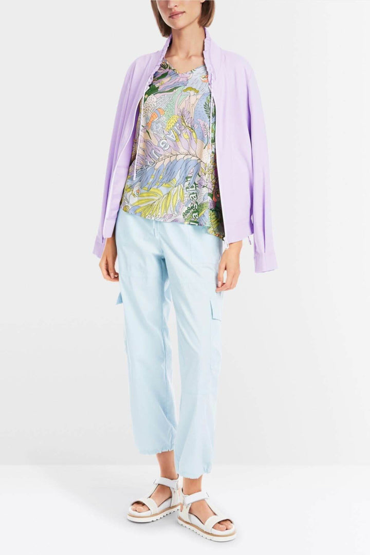 Femme portant une blouse imprimée sushi colorée avec un gilet lilas et un pantalon bleu clair.