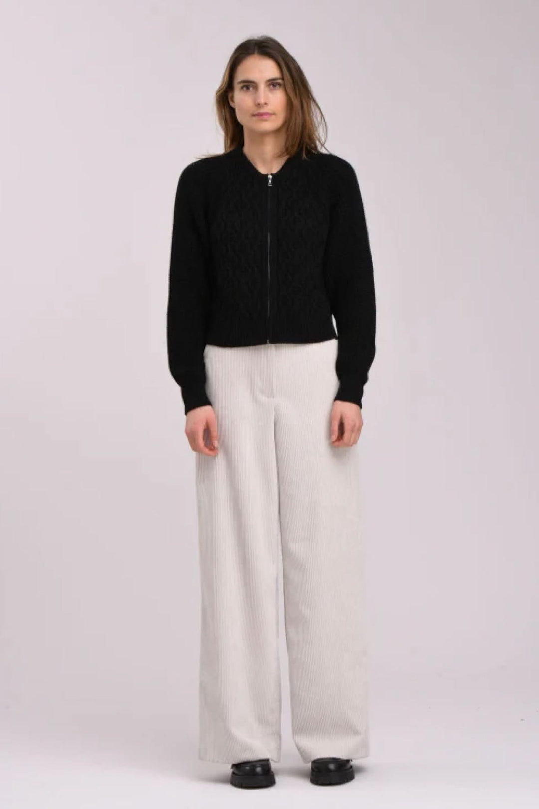 Femme portant un cardigan noir texturé avec fermeture éclair, associé à un pantalon large beige.