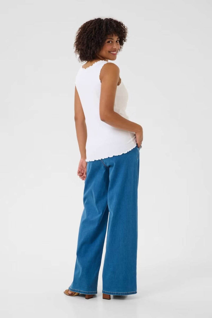 Femme souriante portant une camisole blanche et un pantalon en denim bleu, vue de dos.