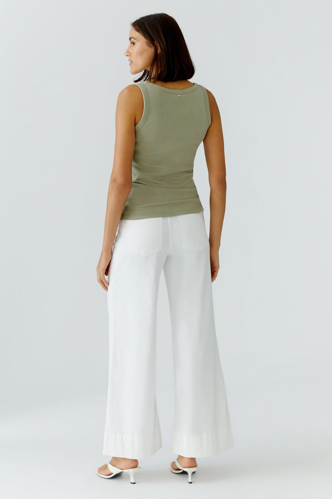 Femme portant une camisole côtelée essentielle verte avec un pantalon blanc, vue de dos. Décris moi le produit en français.