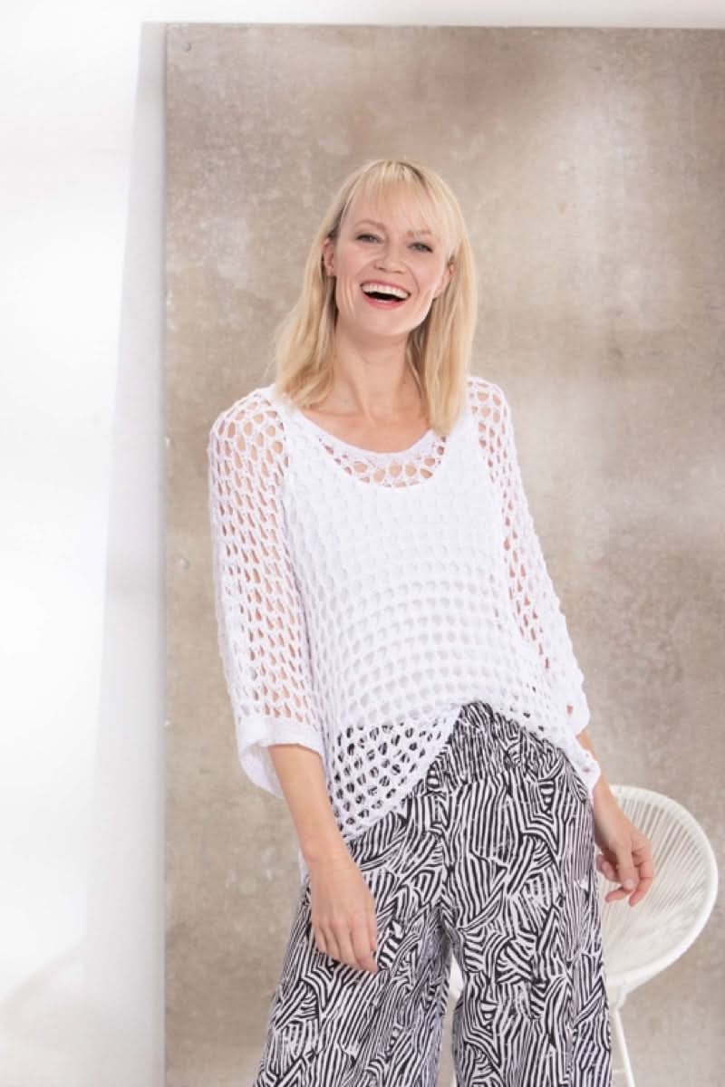 Femme souriante portant un pull blanc crocheté avec un pantalon à motif noir et blanc, fond beige texturé.