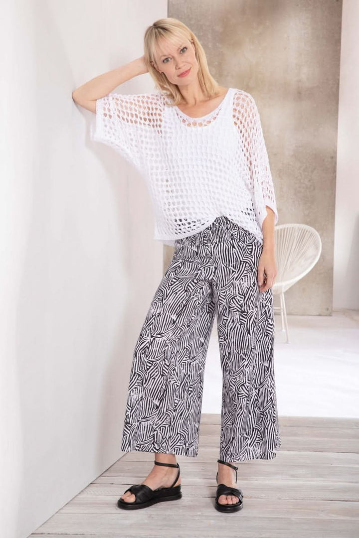 Femme portant un haut en maille blanche et un pantalon large à motifs noirs et blancs, appuyée sur un mur.