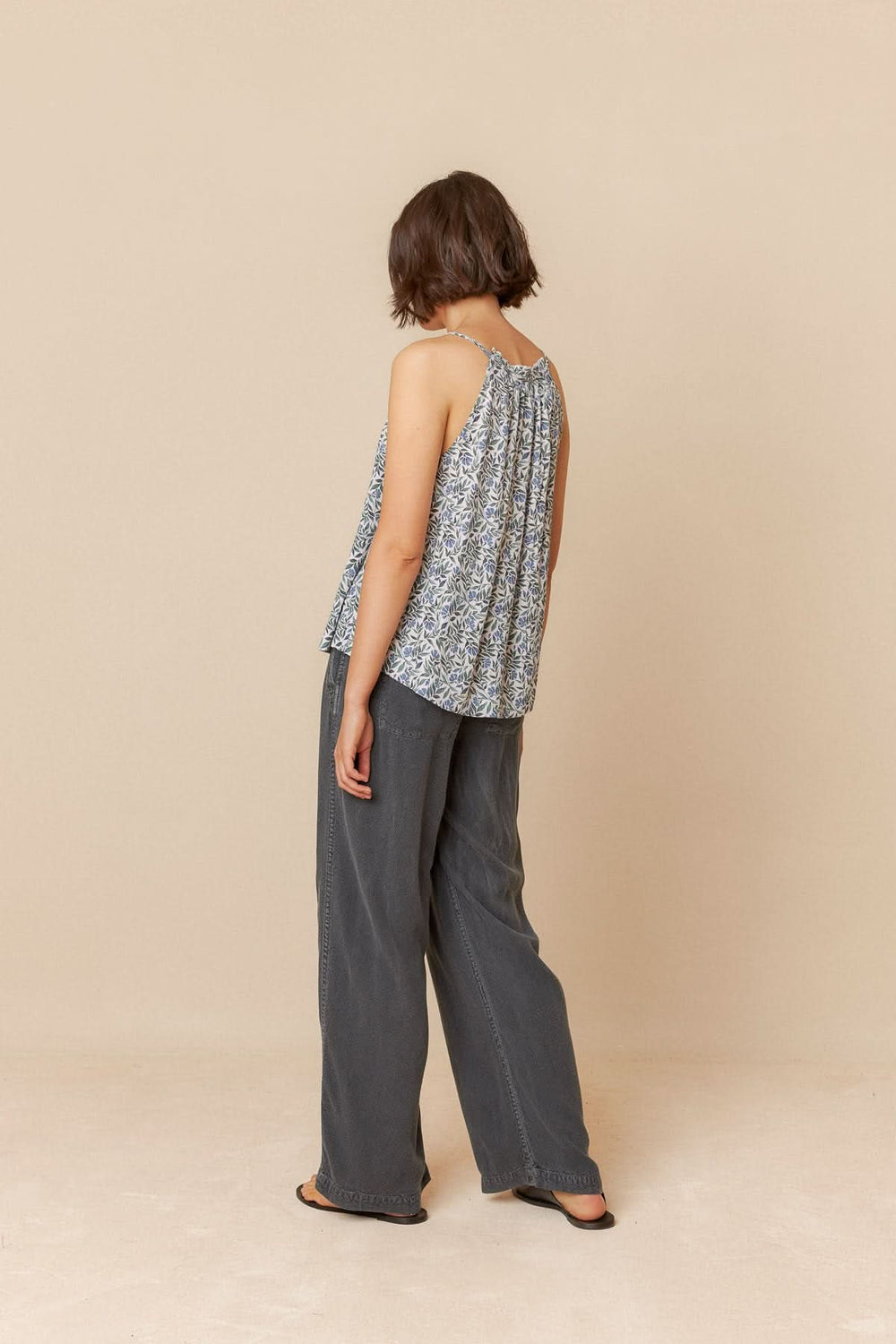 Femme portant une camisole imprimée fleuri sans manches, vue de dos, associée à un pantalon gris.