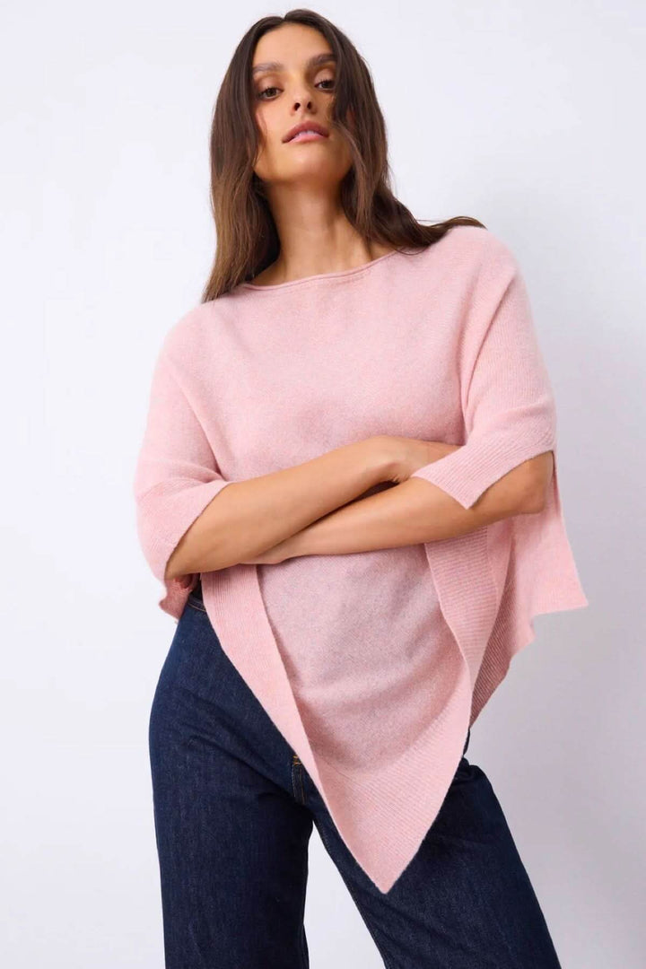 Cape en maille rose à coupe ample et asymétrique, portée par une femme, ajoutant élégance et confort pour superposition en intérieur ou extérieur.