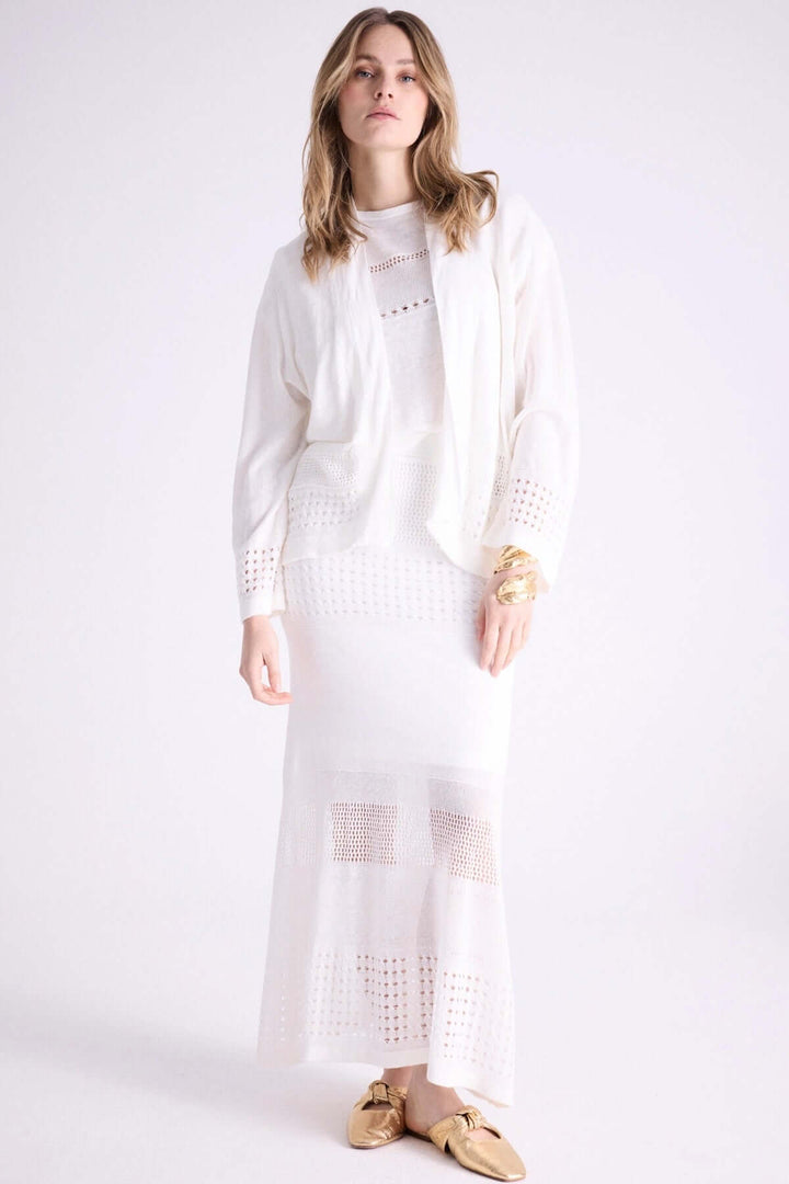 Femme portant un cardigan ouvert blanc avec finitions ajourées, superposé à une robe longue élégante.