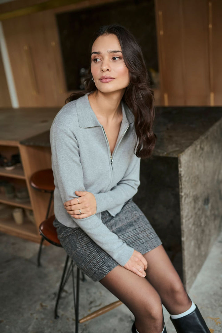 Femme portant un cardigan zippé Visja gris avec col chemise, assise, style minimaliste et moderne, parfait pour un look élégant.