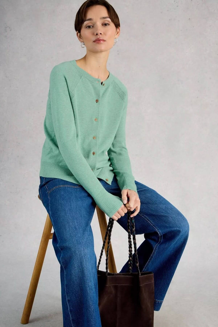 Femme portant un cardigan boutonné en maille légère Lulu vert avec un jean, assise sur un tabouret, tenant un sac noir.