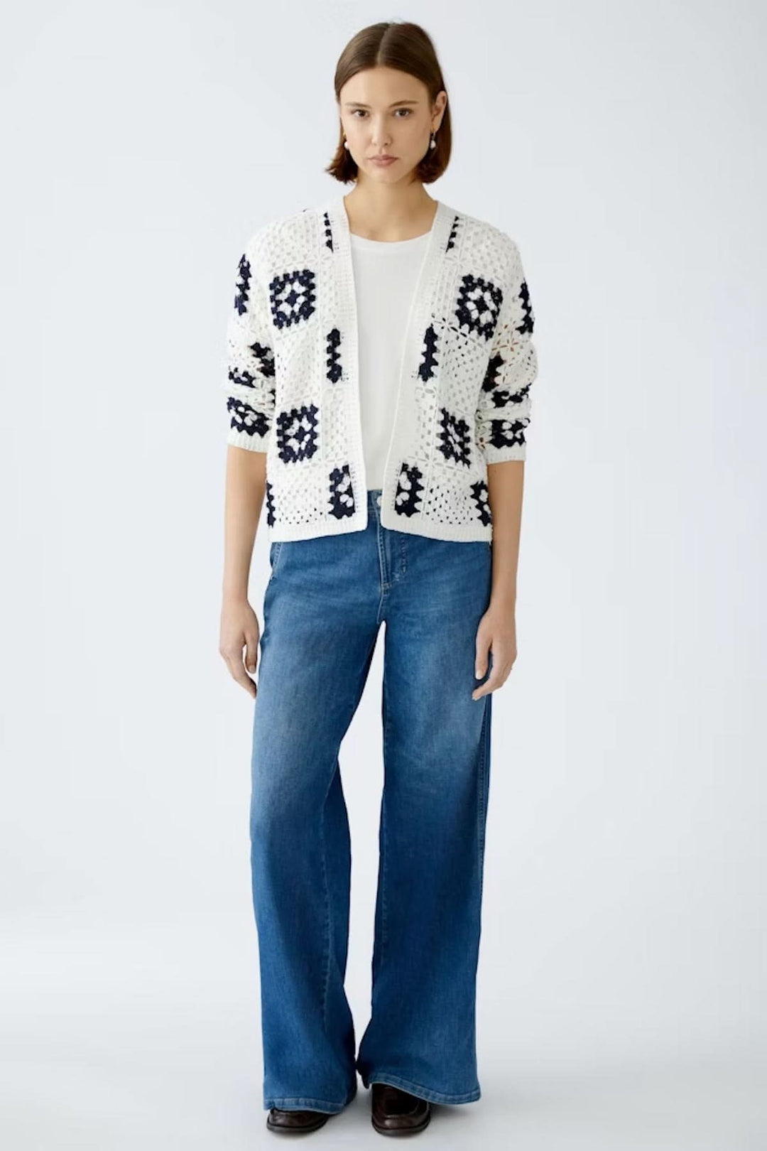 Cardigan en crochet blanc avec motifs géométriques, porté sur un t-shirt blanc et un jean bleu, style bohème.