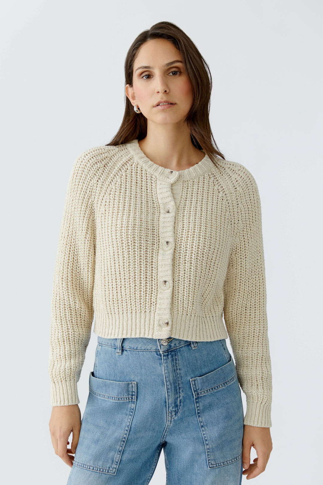 Femme portant un cardigan en maille boutonné crème avec encolure ronde et manches longues, sur un fond gris clair.