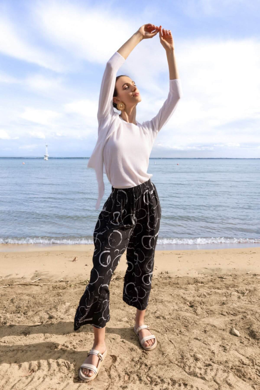 Modèle portant un cardigan léger ouvert blanc sur une plage, associé à un pantalon noir à motifs et des sandales, sous un ciel bleu.