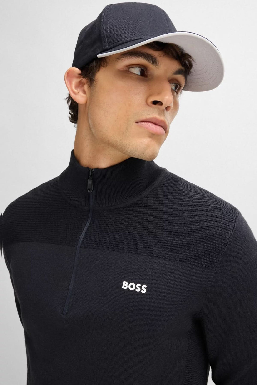 Homme portant une casquette BOSS avec visière et un pull noir à col zippé, affichant un look sportif.