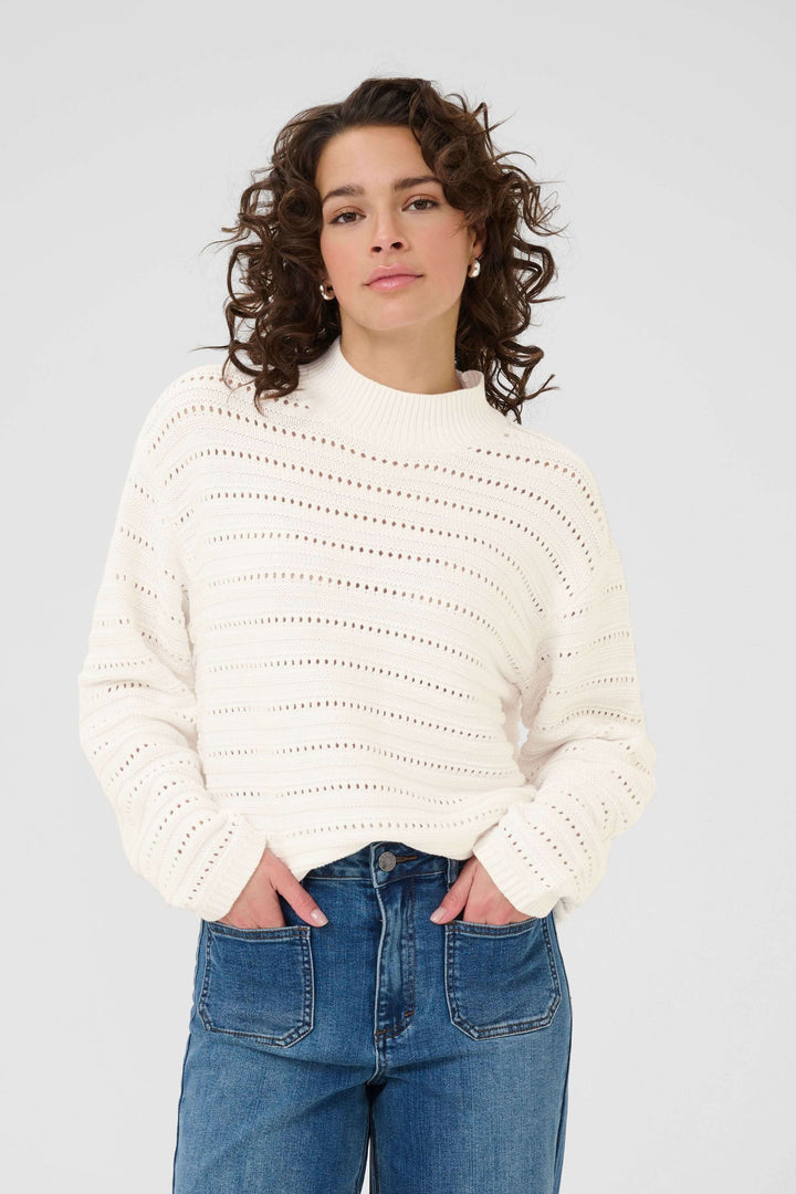 Chandail Dolly en tricot ajouré blanc avec motif subtil, col montant côtelé, et manches longues, porté avec un jean pour un style chic.
