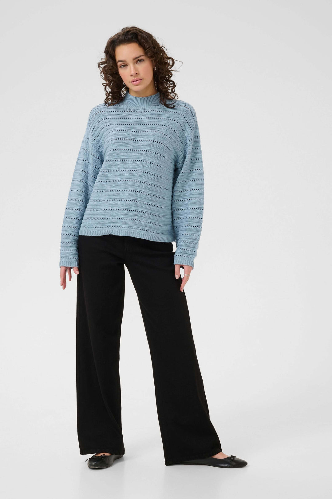 Femme portant le Chandail Dolly bleu avec un motif ajouré, col montant et manches longues, assorti d'un pantalon noir élégant.